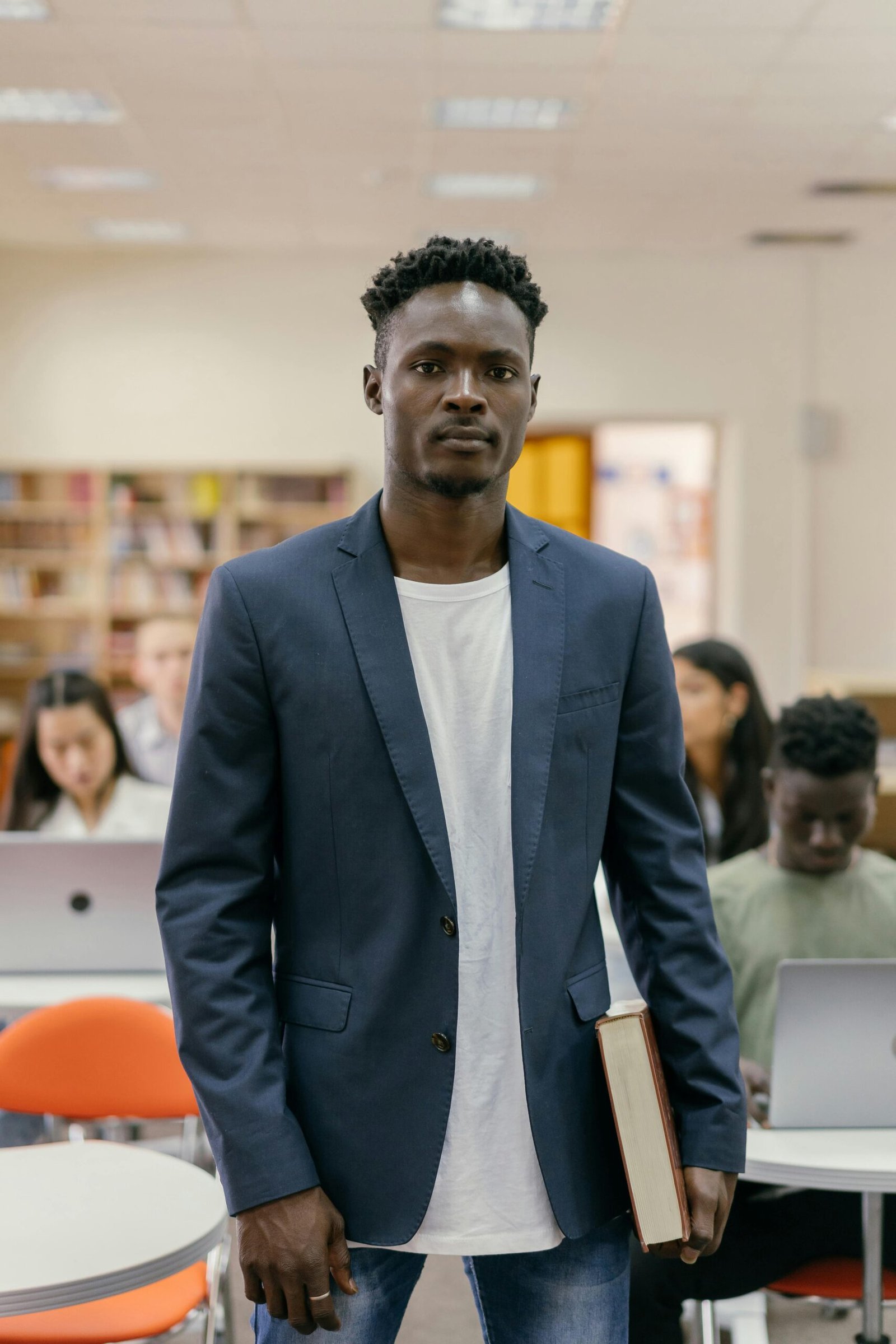 A student stands confidently in a college library surrounded by peers working on laptops.