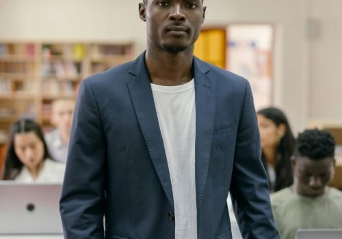 A student stands confidently in a college library surrounded by peers working on laptops.